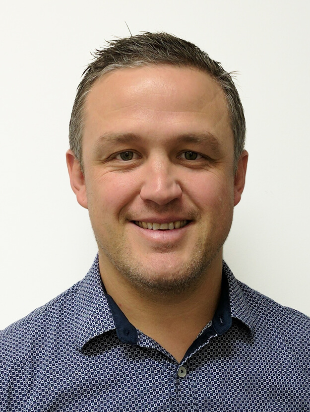 A portrait photo of Geoffrey Norman, Statewide Manager of Lifeline Services Queensland for UnitingCare Queensland, smiling for the camera. Geoffrey has short, sandy coloured hair and stubble on his chin. He wears a blue dotted patterned collared shirt.