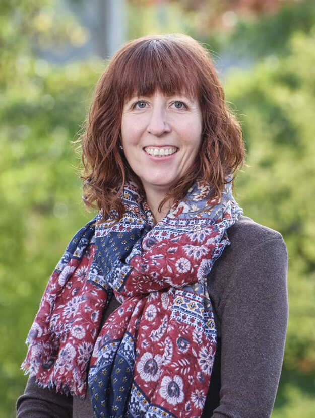 A portrait photo of Nikki Wynne, Director of Services for Wellways Australia, smiling for the camera. Nikki has red shoulder length hair, and wears a large, patterned red and blue scarf around her neck, over a jacket which is a shade of purple.