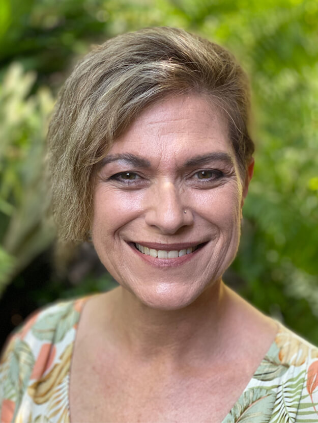 A portrait photo of Sue Fulford, Founding Director of Ther Noldedge House Charity, smiling for the camera. She has a swept-over hairstyle, and wears a top with a print of green and orange leaves.
