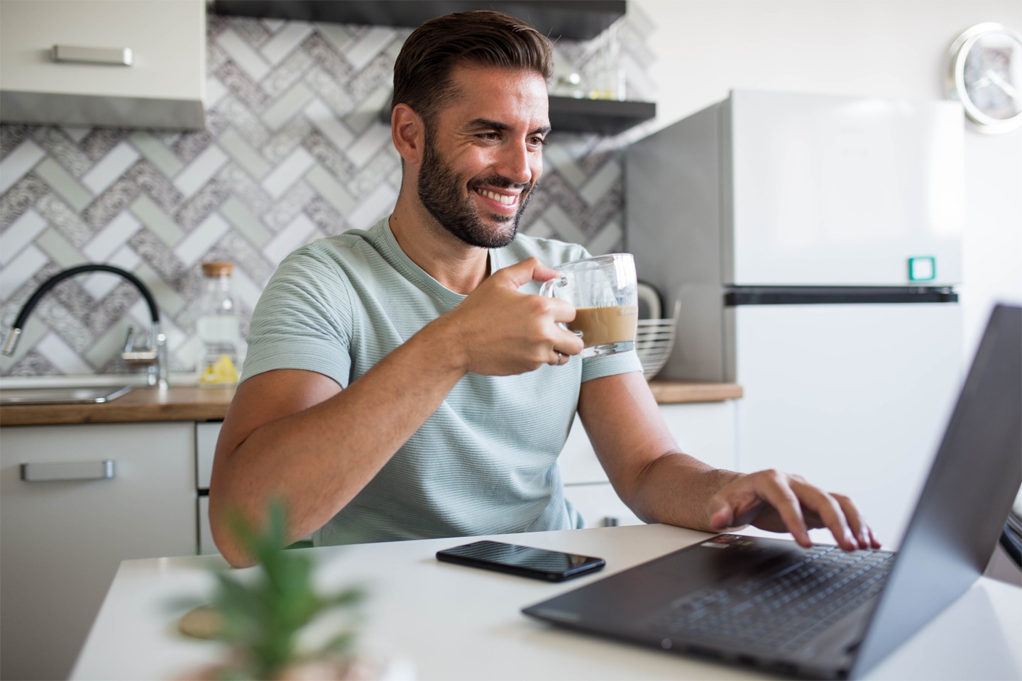 A man working from home, sitting at a desk, looking at his laptop, holds a cup. He is smiling